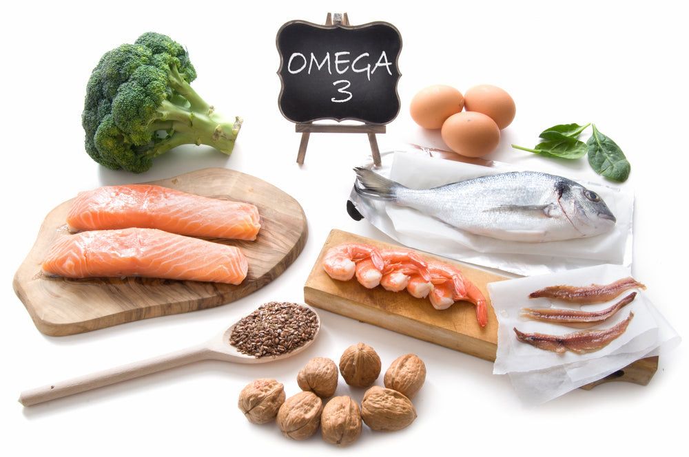 A variety of foods rich in omega-3 fats, including salmon, shrimp, eggs, broccoli, walnuts, and flaxseeds, are arranged around a chalkboard labeled