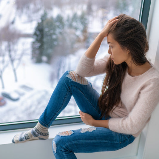 A young woman sits by a window, resting her head on her hand with a thoughtful expression, while looking out at a snowy landscape and trees in the background.