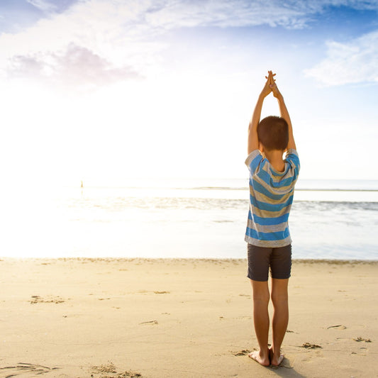 A child in a striped shirt stands on a sandy beach, arms raised towards the bright sky, with the ocean gently lapping at the shore in the background.