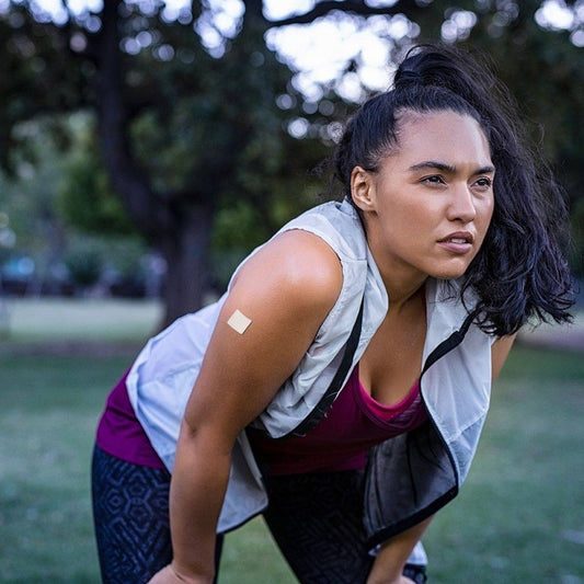 A woman in athletic wear leans forward with a focused expression while exercising outdoors, with a vitamin patch visible on her arm, surrounded by greenery in a park.