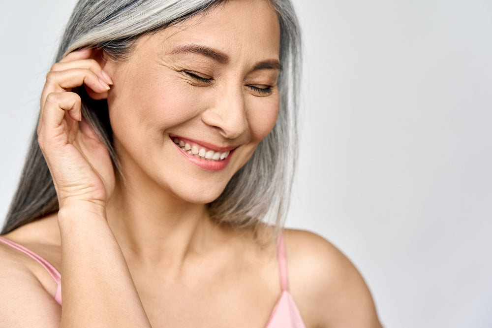 A smiling older woman with long, gray hair gently touches her ear while closing her eyes, conveying a sense of joy and contentment against a soft, neutral background.