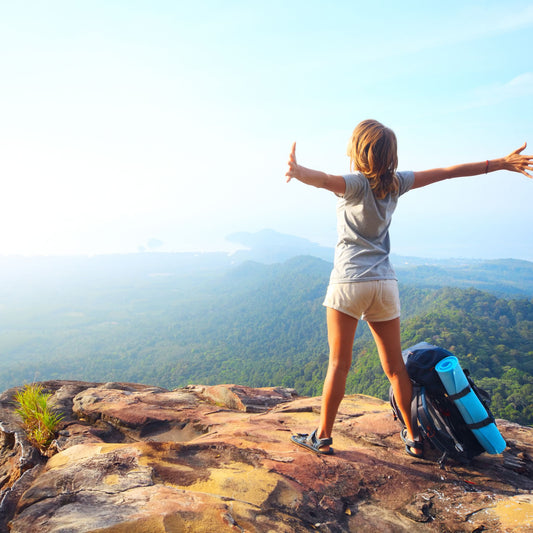 A person stands on a rocky ledge with arms outstretched, overlooking a lush green landscape and distant hills, symbolizing energy and vitality, possibly linked to the benefits of Vitamin B12.