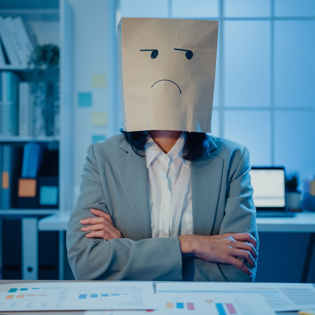 A person in a gray blazer sits at a desk with crossed arms, wearing a paper bag with a sad face drawn on it, in a dimly lit office with charts and a computer in the background.
