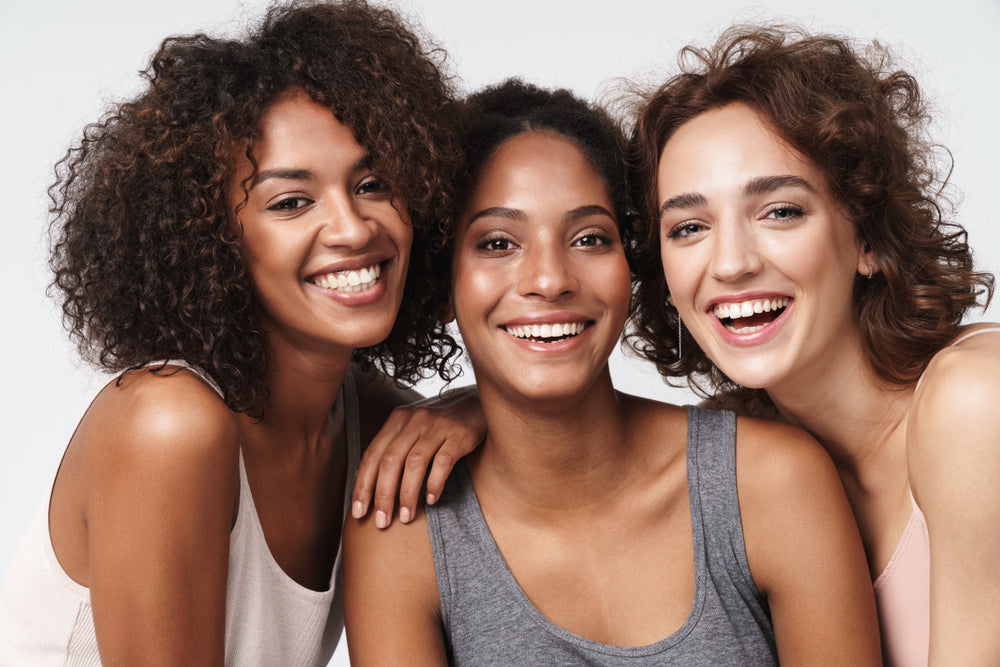 Three women with curly hair smile joyfully while standing close together against a light background, showcasing healthy complexions and a sense of friendship.