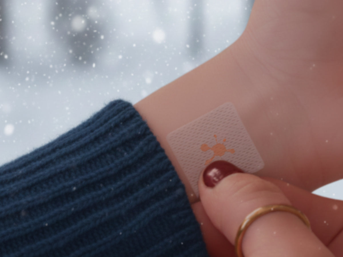 Person wearing a cozy blue sweater applying a small vitamin patch to their wrist on a snowy winter day.