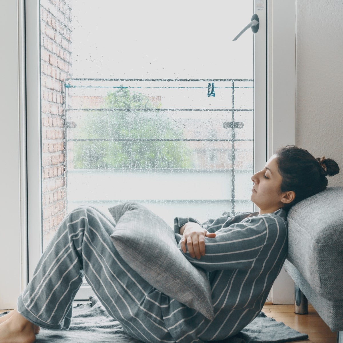 A woman in striped pajamas sits curled up on a floor mat, resting her head against a couch while gazing out a rain-streaked window, conveying a sense of solitude and introspection.