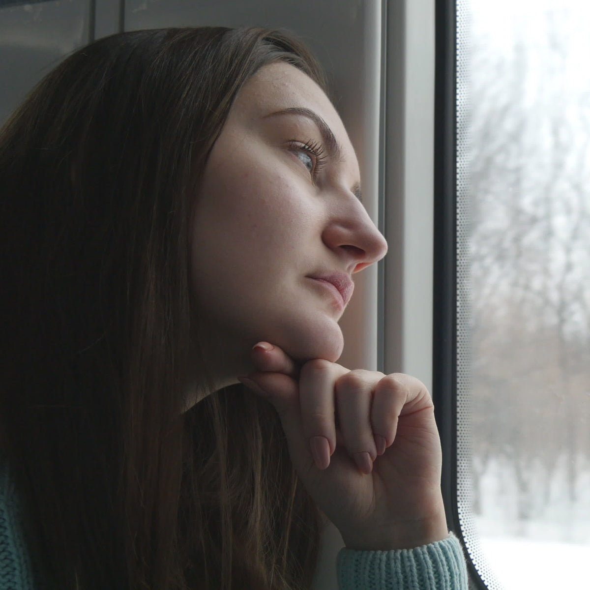 A young woman rests her chin on her hand, gazing thoughtfully out a window at a snowy landscape, reflecting feelings associated with seasonal affective disorder.