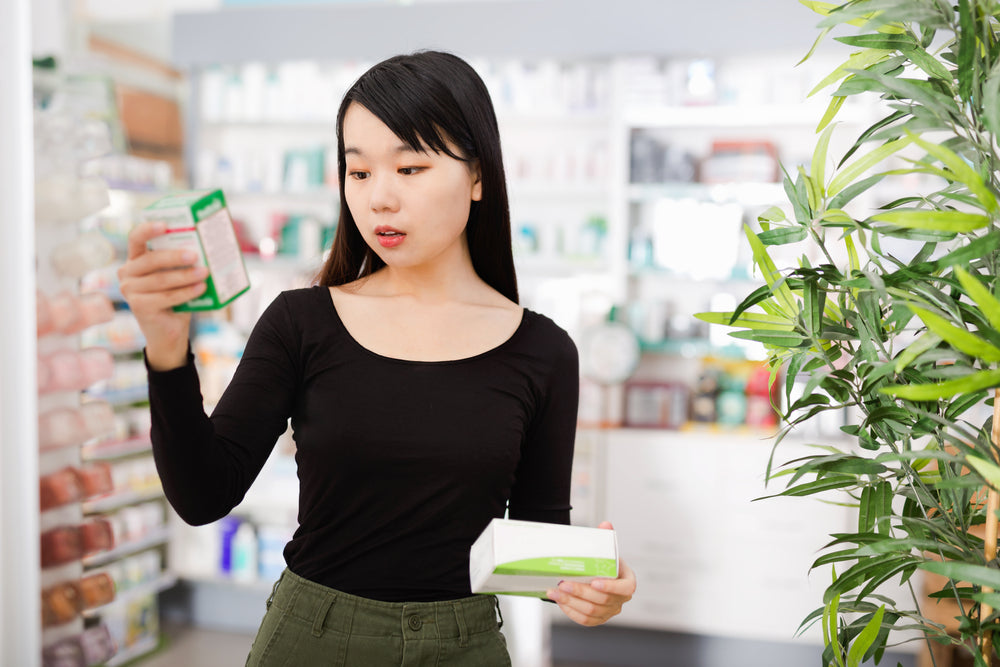 A young woman in a black long-sleeve shirt examines two vitamin products in a pharmacy, comparing their packaging while surrounded by shelves of health and beauty items.