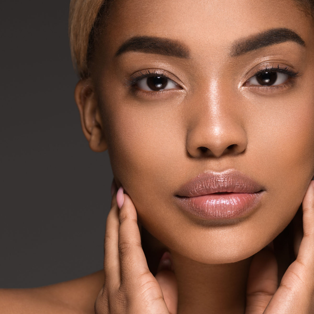 A close-up of a young woman with smooth skin gently touching her face, showcasing a natural, radiant look against a dark background, highlighting the theme of skincare and beauty.