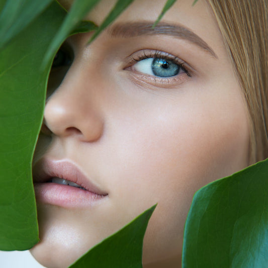 A close-up of a young woman’s face partially obscured by green leaves, showcasing her striking blue eye and soft expression against a natural background.