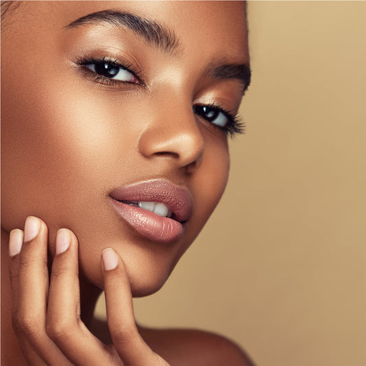 A young woman with glowing skin gently touches her chin, smiling softly against a warm beige background, highlighting tips for achieving clear skin.