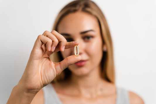 A woman with long hair holds a collagen supplement capsule up to the camera, looking thoughtfully at it against a plain white background.