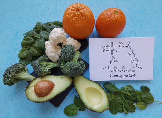 A variety of fresh vegetables, including broccoli, cauliflower, spinach, and avocado, along with two oranges and a card displaying the chemical structure of Coenzyme Q10, are arranged on a blue background.