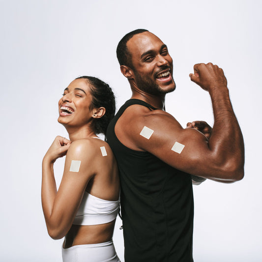 A smiling man and woman stand back-to-back, flexing their arms, each with a vitamin patch on their shoulders, against a plain white background.
