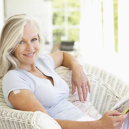 A smiling woman with long, light hair sits comfortably in a wicker chair, holding a magazine while enjoying a bright, airy room filled with natural light.