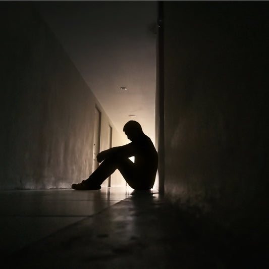 A silhouetted figure sits on the floor of a dimly lit hallway, hugging their knees and appearing contemplative, suggesting a moment of solitude and reflection.