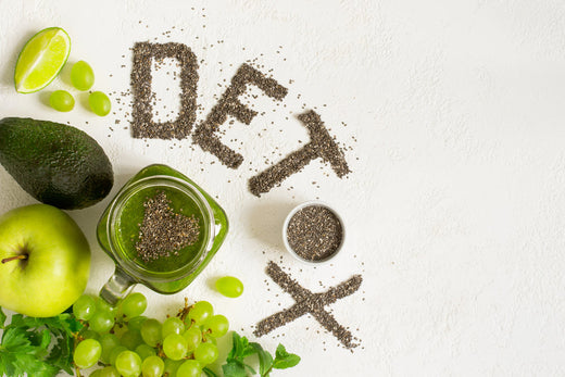 Green fruits and vegetables, including an avocado, green apple, and grapes, surround a jar of green smoothie, with the word "DETOX" creatively spelled out in chia seeds on a light surface.