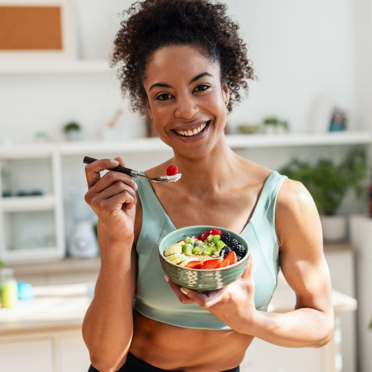 A smiling woman in a light green tank top holds a bowl of colorful fruit and yogurt while preparing to take a bite with a fork, standing in a bright kitchen filled with plants and light.