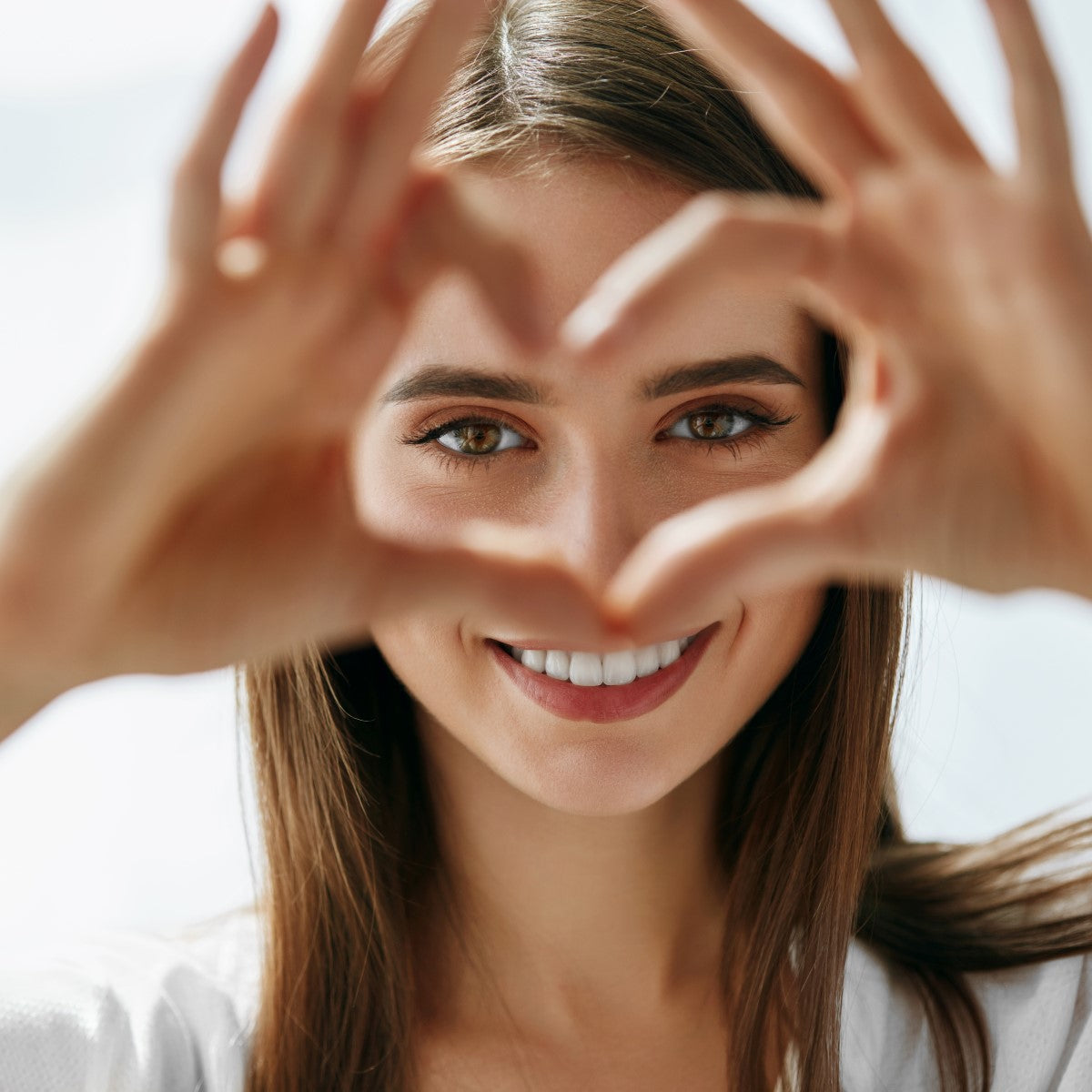 A young woman smiles brightly while forming a heart shape with her hands, set against a soft, light background that suggests a cheerful atmosphere.