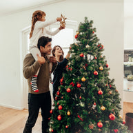 A man supports a girl on his shoulders as she places a star on top of a decorated Christmas tree, while a woman smiles and adds ornaments, all in a bright, cozy living room.