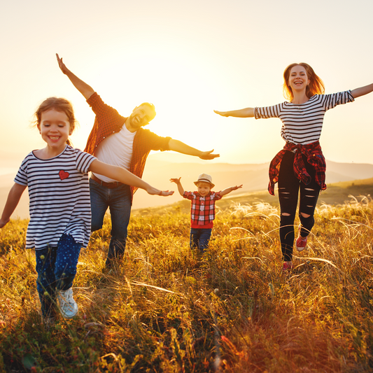 A joyful family of four, including two children, runs through a sunlit field, with arms outstretched and smiles on their faces, enjoying a vibrant sunset in the background.