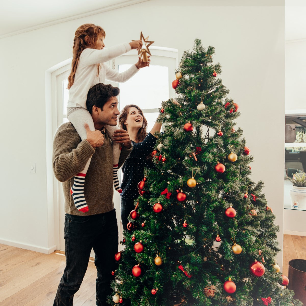 A man supports a girl on his shoulders as she places a star on top of a decorated Christmas tree, while a woman smiles and adds ornaments, all in a bright, cozy living room.