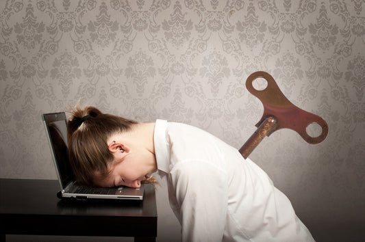 A woman in a white shirt rests her head on a laptop while a large wind-up key protrudes from her back, set against a patterned wallpaper background, illustrating exhaustion and the need for nutrient absorption.