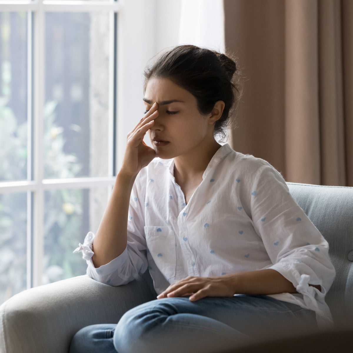 A young woman sits on a couch, holding her forehead with a pained expression, in a well-lit room with large windows, suggesting she may be experiencing discomfort related to nutritional deficiency.