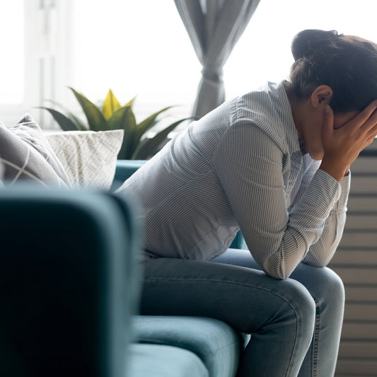 A woman sits on a teal couch with her head in her hands, looking distressed, in a bright room with plants and curtains, reflecting the emotional impact of vitamin deficiency.