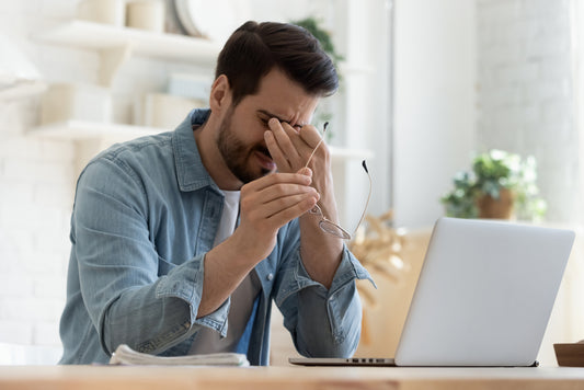 A man in a denim jacket sits at a desk, rubbing his eyes in frustration while holding his glasses, with a laptop and plants visible in a bright, modern room.