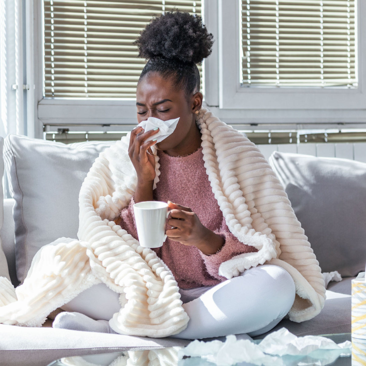 A woman wrapped in a cozy blanket sits on a couch, holding a cup while blowing her nose with a tissue, surrounded by crumpled tissues and natural light from nearby windows.