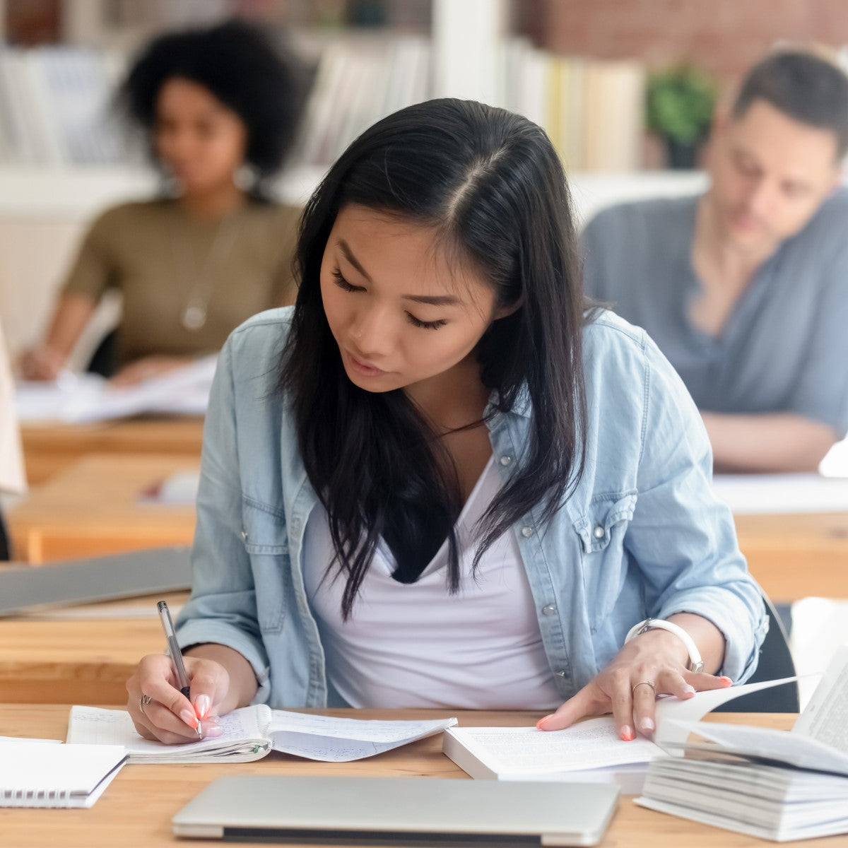 A student with long dark hair focuses intently on writing in a notebook while surrounded by books and classmates in a bright, modern study space.