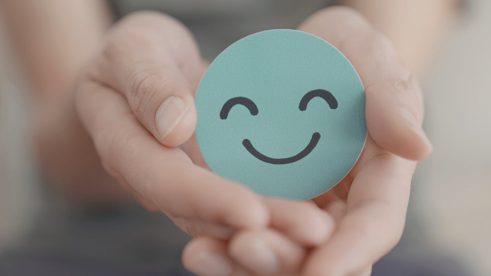 A person holds a round, teal card with a smiling face in their hands, conveying a message of positivity and well-being in a soft, neutral setting.