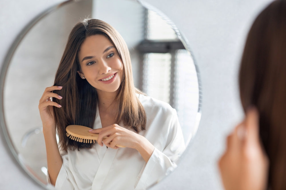 A woman in a white robe smiles while brushing her long, straight hair in front of a round mirror, with soft natural light illuminating the modern bathroom setting.