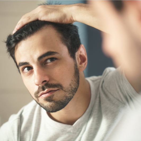 A man with short dark hair examines his scalp in a mirror, looking concerned while running his hand through his hair, suggesting a focus on hair health or thinning.