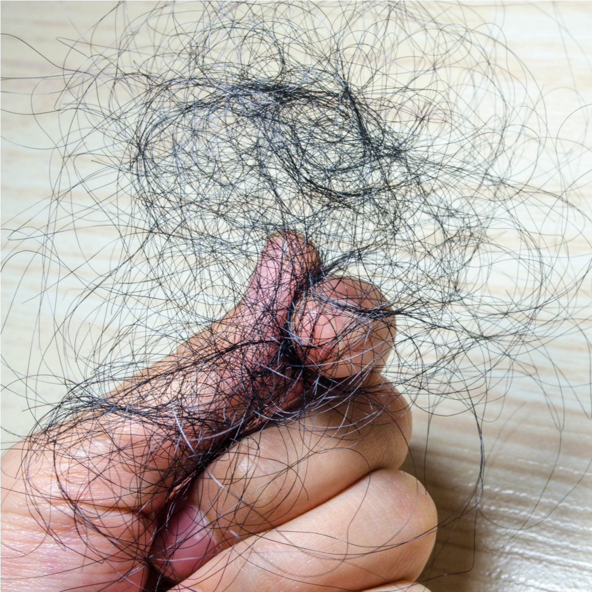 A hand holds a tangled mass of dark hair strands against a light wooden surface, illustrating the issue of temporary hair loss.