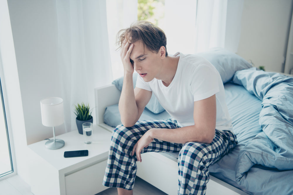 A young man in pajamas sits on the edge of a bed, holding his head with one hand and looking distressed, in a bright room with a glass of water and a phone nearby, suggesting a hangover.