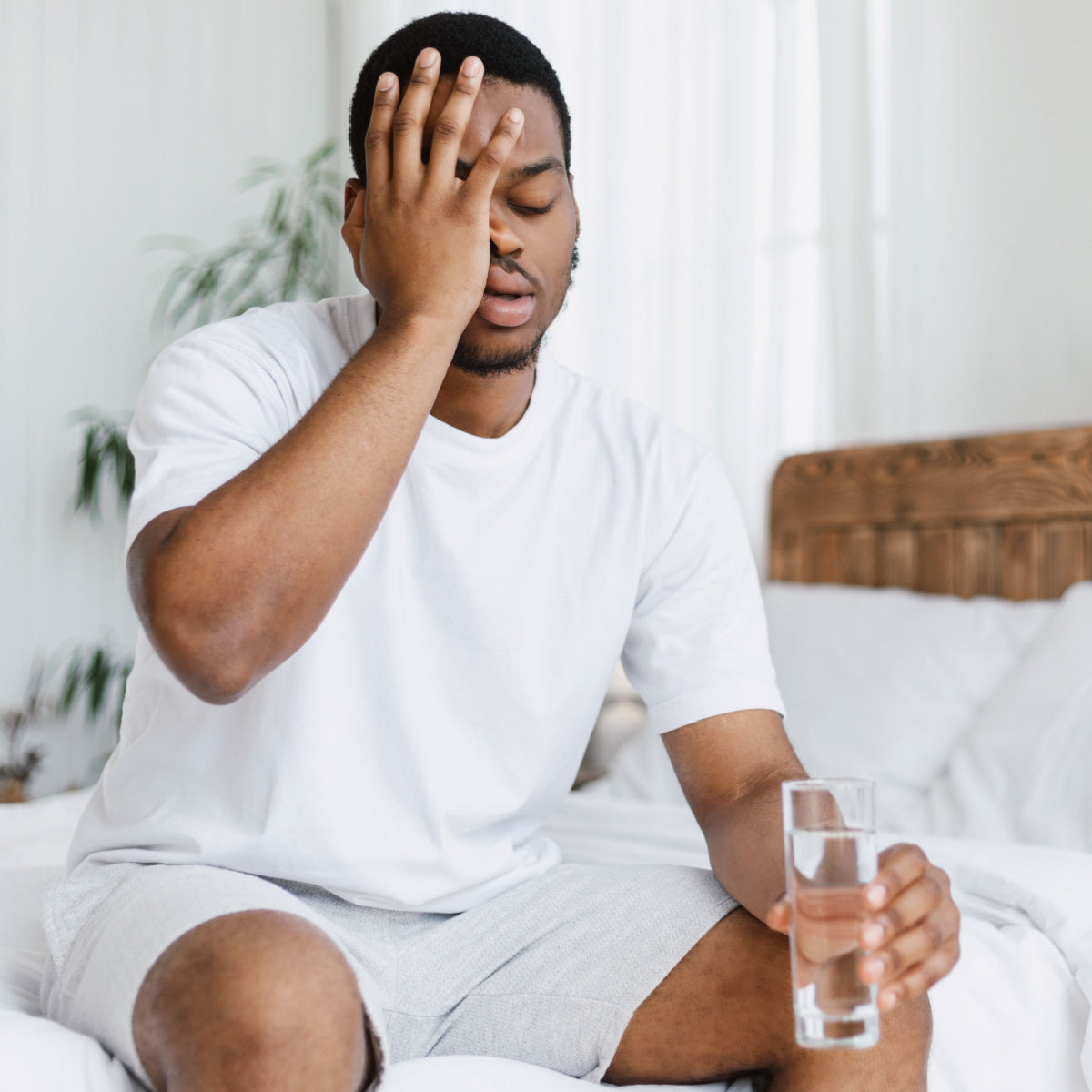 A man sits on a bed, holding a glass of water and resting his forehead on his hand, appearing tired and distressed, likely recovering from a hangover in a bright, airy room.