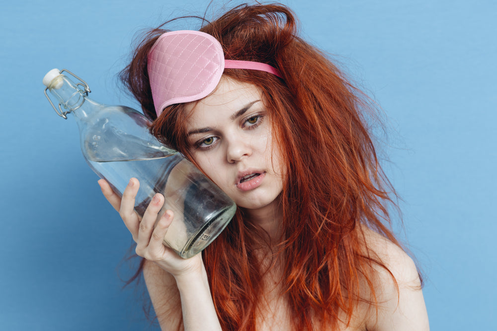 A disheveled young woman with red hair, wearing a pink sleep mask, leans her head against a glass bottle of water, looking tired against a light blue background, suggesting a struggle with a hangover.