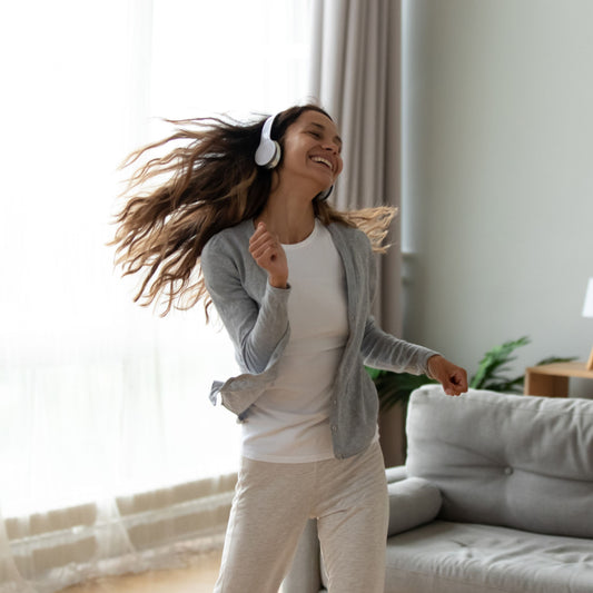 A woman with long hair dances joyfully in a cozy living room while wearing headphones, dressed in a light gray cardigan and white shirt, surrounded by natural light and a comfortable sofa.