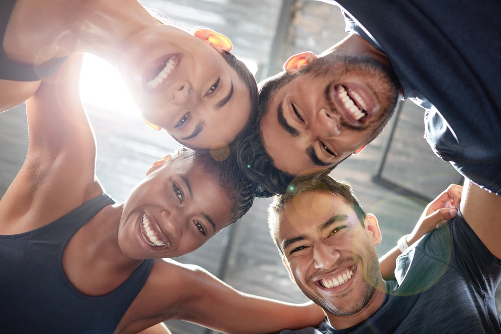 Four smiling friends lean in together in a circle, showing joy and connection, with bright light streaming in from above, suggesting a positive atmosphere related to health and wellness.