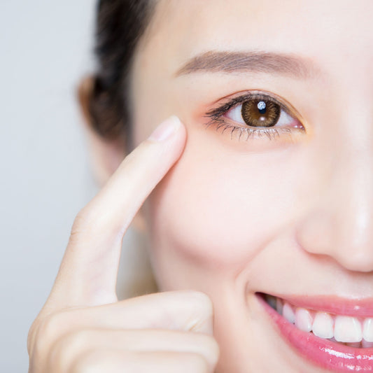 A smiling woman points to her eye, demonstrating a tip for maintaining healthy eyes, against a light gray background.