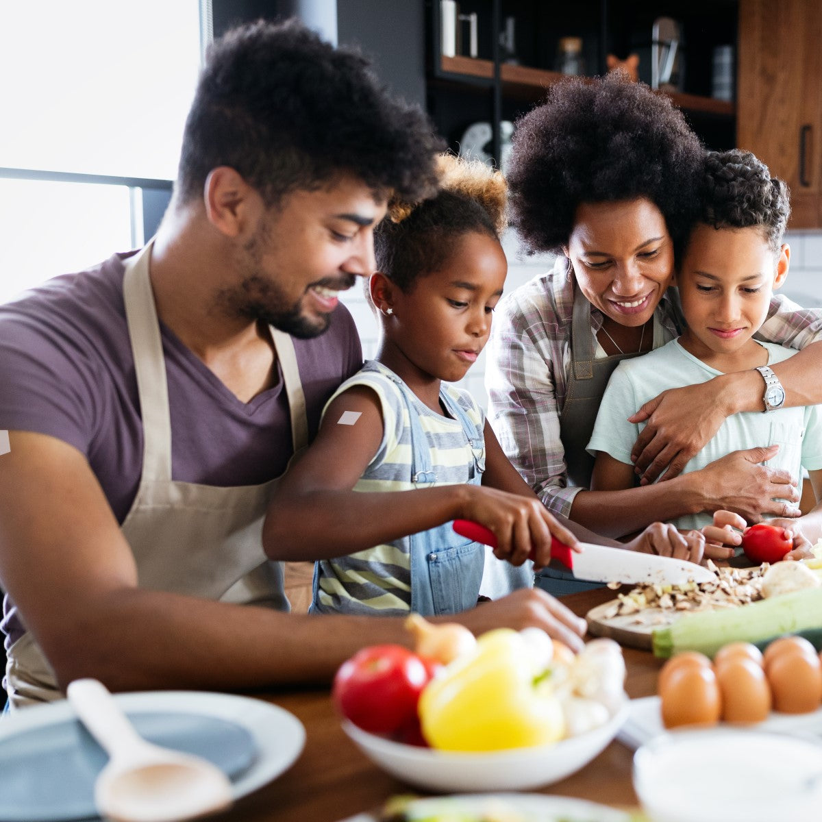 A family of four, including two children, joyfully prepares food together in a modern kitchen, with one child chopping vegetables while the parents smile and embrace the other child.
