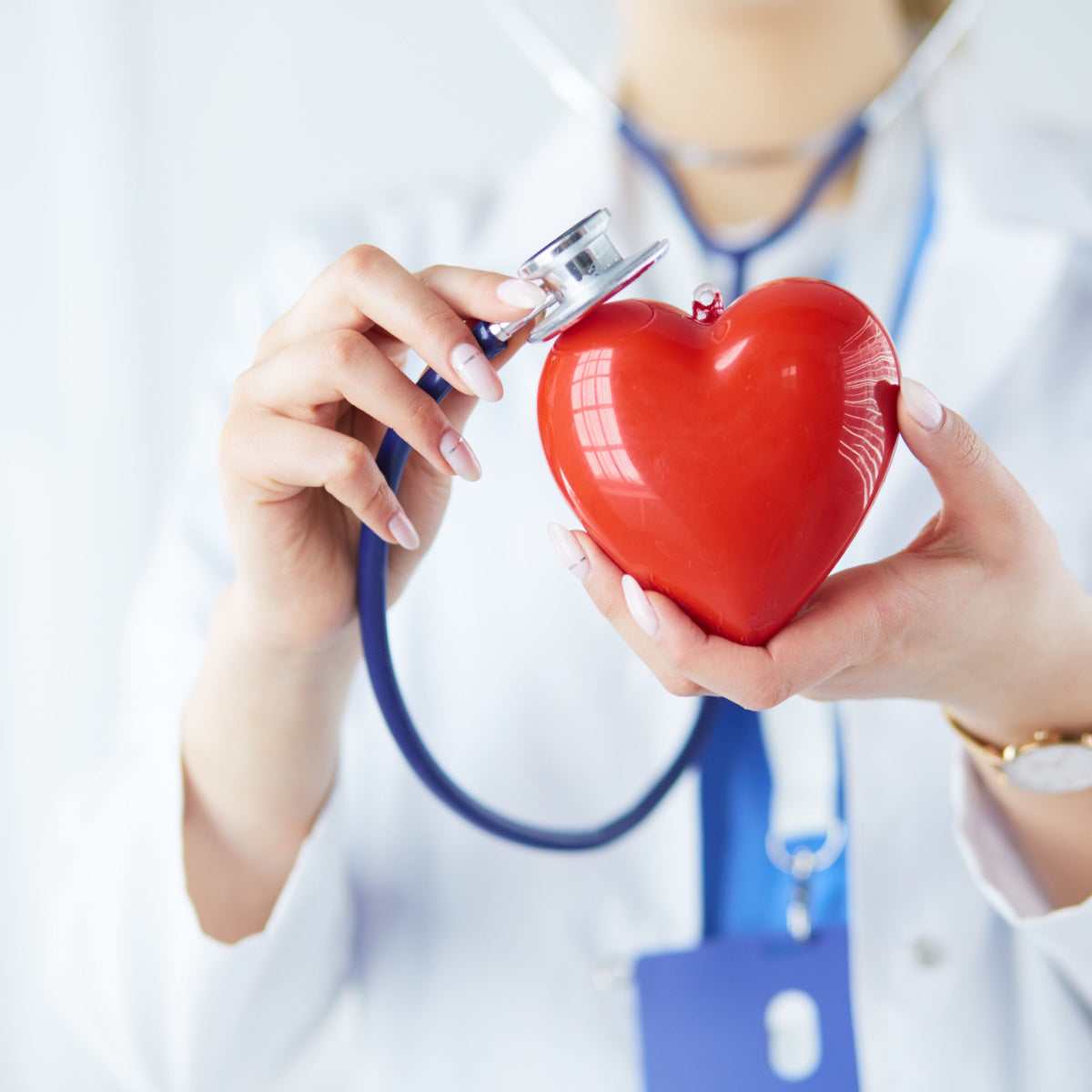 A healthcare professional in a white coat holds a red heart model while placing a stethoscope on it, symbolizing heart health and wellness in a bright, clinical setting.