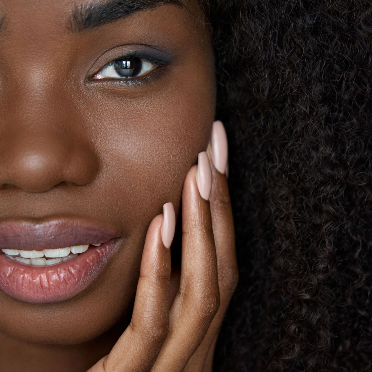 A woman with curly hair gently touches her cheek, showcasing her smooth skin and well-groomed nails, while smiling softly at the camera in a close-up portrait.