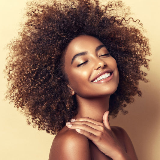 A young woman with curly hair smiles brightly while gently touching her shoulder, set against a soft, neutral background, highlighting her radiant skin and joyful expression.