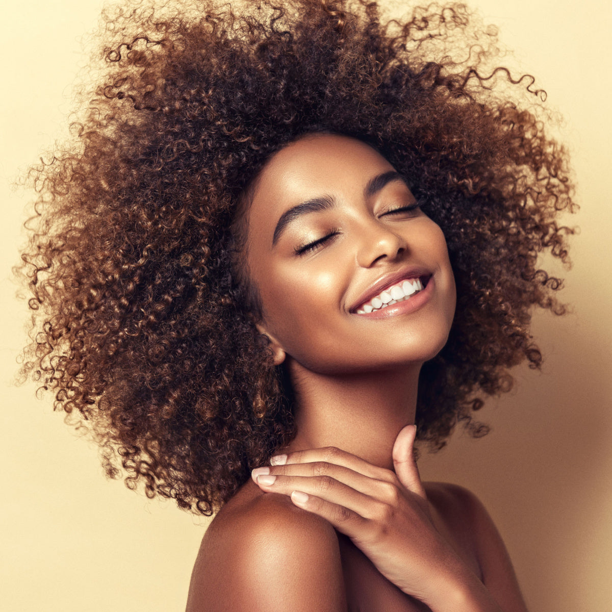 A young woman with curly hair smiles brightly while gently touching her shoulder, set against a soft, neutral background, highlighting her radiant skin and joyful expression.