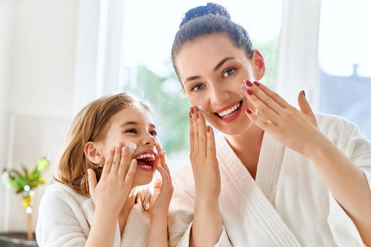 A woman and a young girl, both in white bathrobes, smile and apply skincare products to their faces in a bright, sunny bathroom, sharing a joyful moment together.
