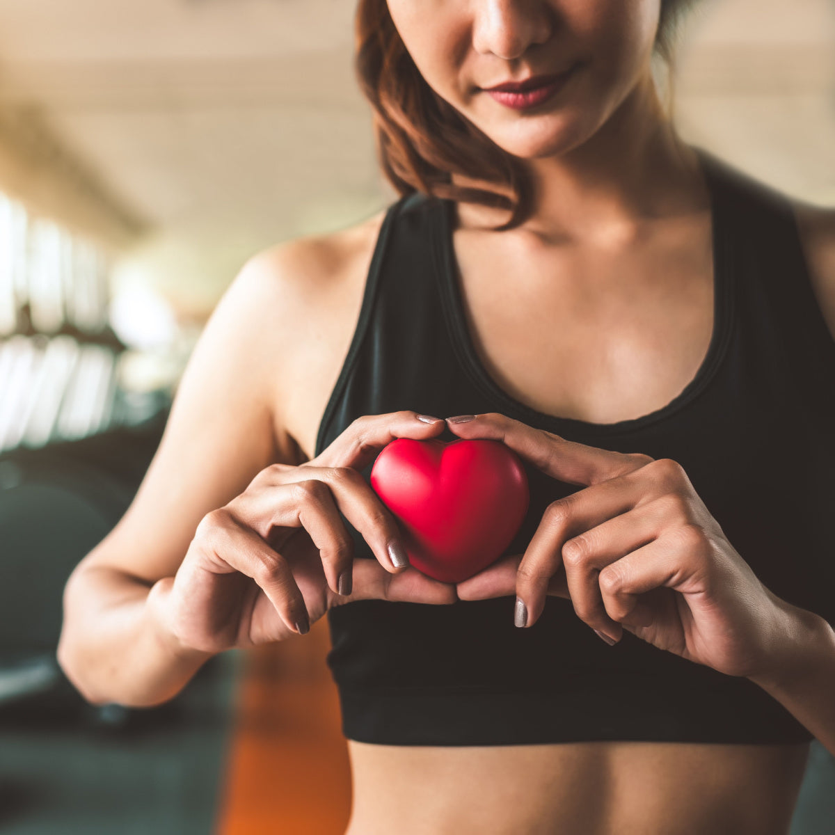 A woman in a black sports top holds a small red heart symbol in front of her chest, conveying a message about heart health in a bright gym setting.