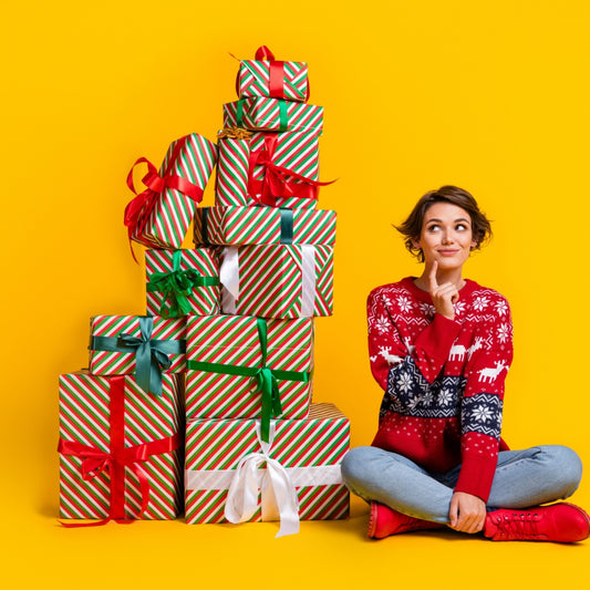 A woman in a festive red sweater sits cross-legged on the floor, thoughtfully smiling while surrounded by a stack of colorful holiday gift boxes against a bright yellow background.